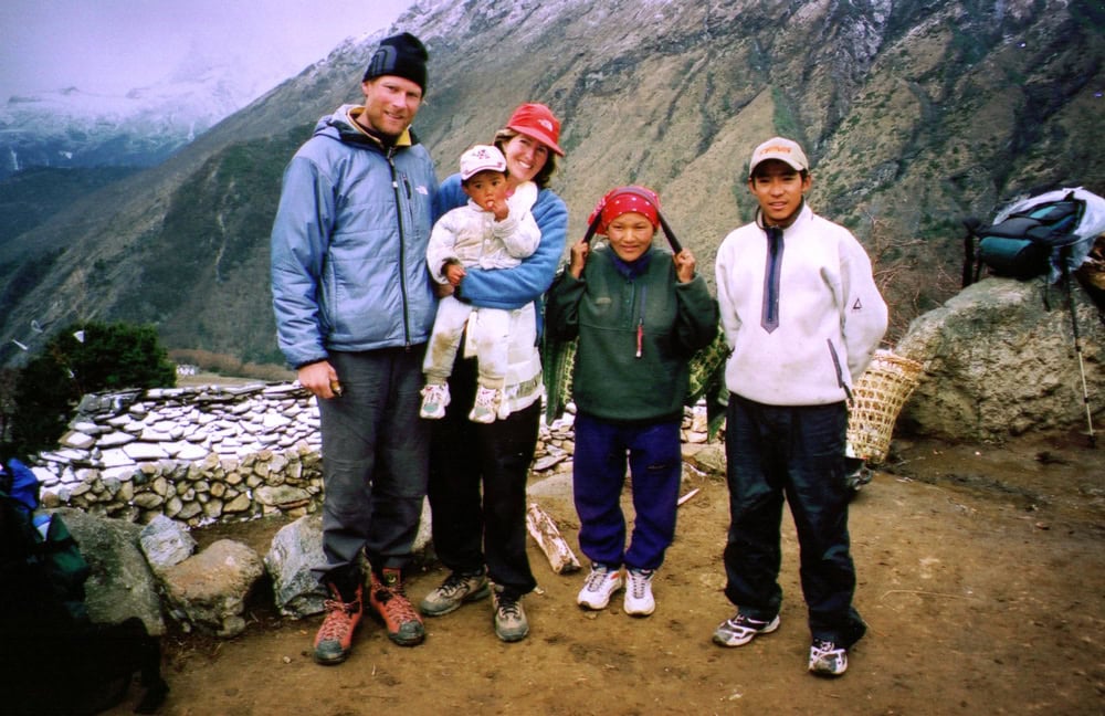 Two adults, one holding a child, pose with three locals in a mountainous area of Nepal. One local shows a peace sign with both hands. Snow-capped mountains and rocky terrain are visible in the background, perfect for a family photobook memory.