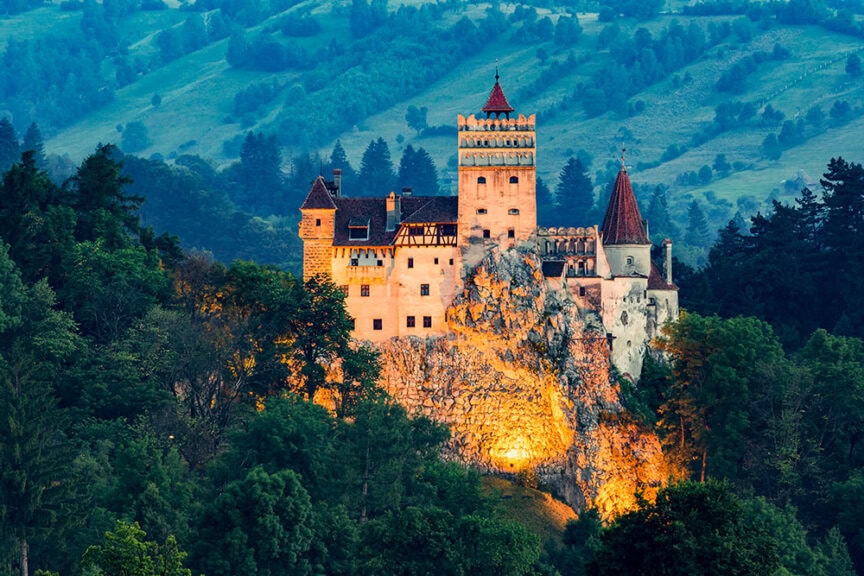 A historic citadel sits on a rocky hill surrounded by dense forest and green hills, with warm lights illuminating its stone structure.