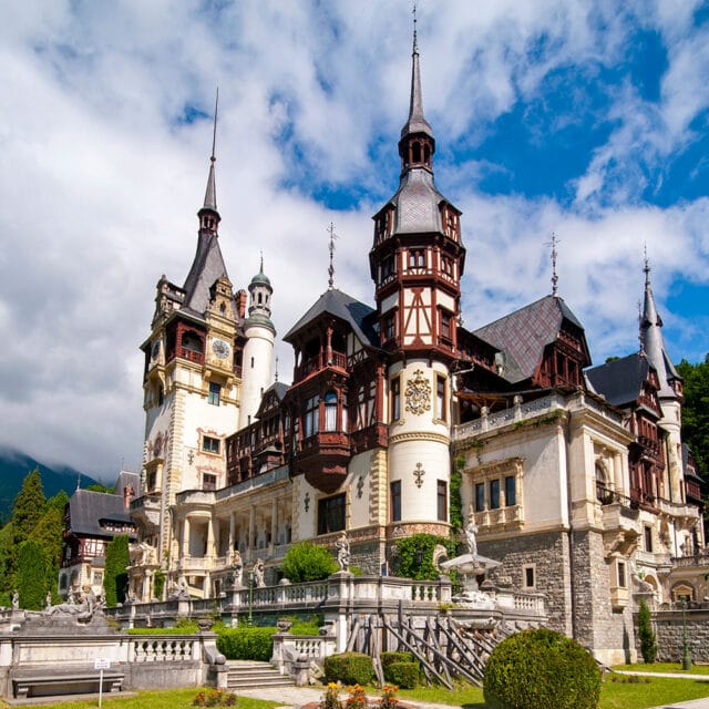 A grand, ornate castle with multiple spires and intricate detailing stands against a backdrop of trees and a partly cloudy sky, surrounded by well-manicured gardens and statues, evoking the majestic allure of ancient citadels.