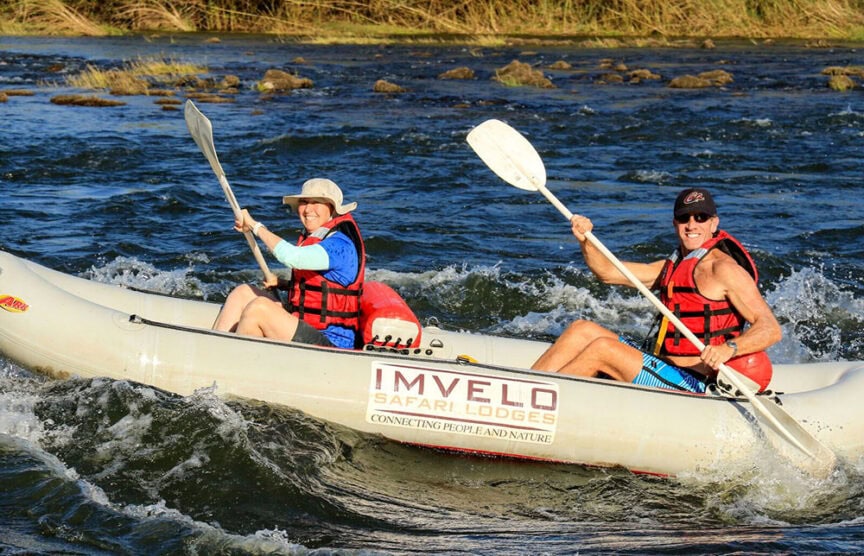 Two people in life jackets paddle a raft labeled "Imvelo Safari Lodges" through whitewater rapids in amazing morning light, capturing the adventurous spirit of Zimbabwe.