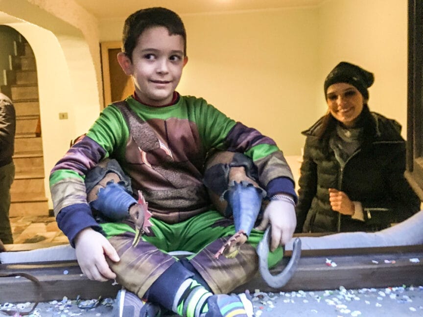 A child dressed in a colorful costume sits on a windowsill, with a woman in a black jacket and hat nearby, reminiscent of the vibrant Pagan Festivals in Sardinia. Confetti is scattered around.