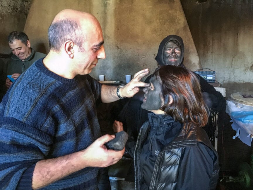 A man applies black paint to a woman's face, while another person with a painted face looks on. In the background, someone appears to be smiling. The scene evokes the rich Sardinian traditions often seen during pagan festivals in Sardinia. Indoor, casual setting.
