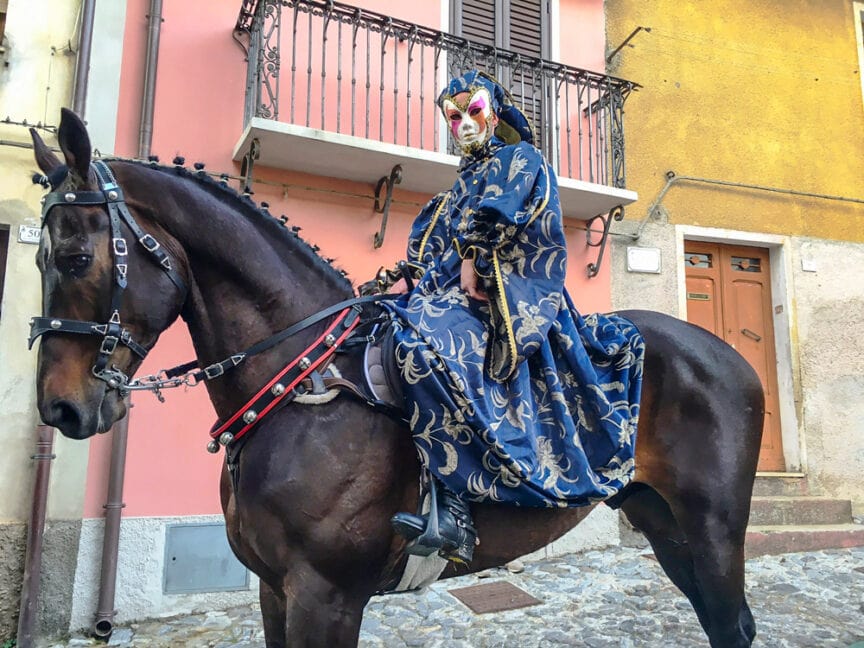 A person wearing a masquerade costume and mask is riding a dark horse on a cobblestone street, with colorful buildings in the background, as if parading through the vibrant traditions of Sardinia's Pagan Festivals.
