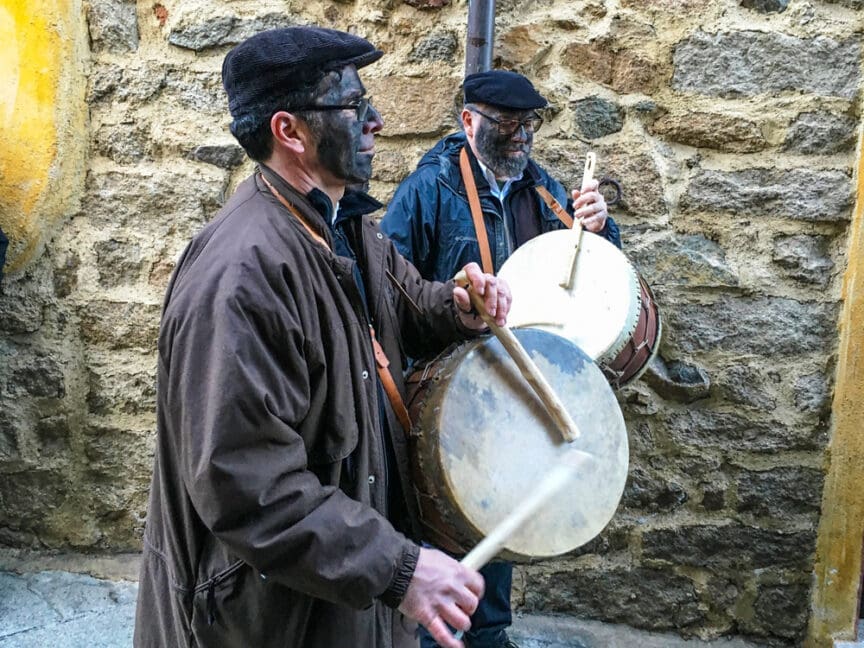 Two men with painted faces play drums on a cobblestone street in Sardinia, against a stone wall, dressed in brown and black jackets with black hats. The rhythmic beats are reminiscent of ancient Pagan Festivals, echoing through the historic town.