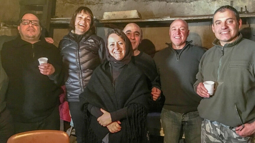 A group of six people, four men and two women, standing closely together indoors in a dimly-lit, rustic room in Sardinia, smiling at the camera. Some are holding small white cups, perhaps sharing stories from local Pagan festivals.