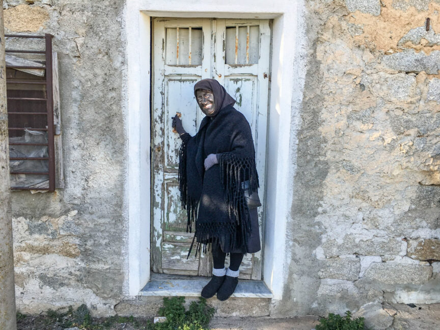 A person dressed in black stands in front of an old, weathered door and stone wall in Sardinia, holding the door handle, perhaps recalling ancient Pagan Festivals.