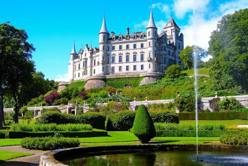 A large medieval-style citadel with multiple turrets sits on a green hillside, with manicured gardens and a fountain in the foreground.