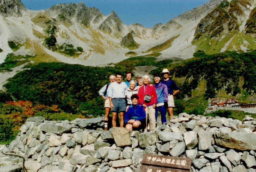 A group of eight people in outdoor attire pose for a photo on a stone structure with a mountainous backdrop, capturing their journey. A sign in the foreground has text in Japanese, reminiscent of an ancient haiku.