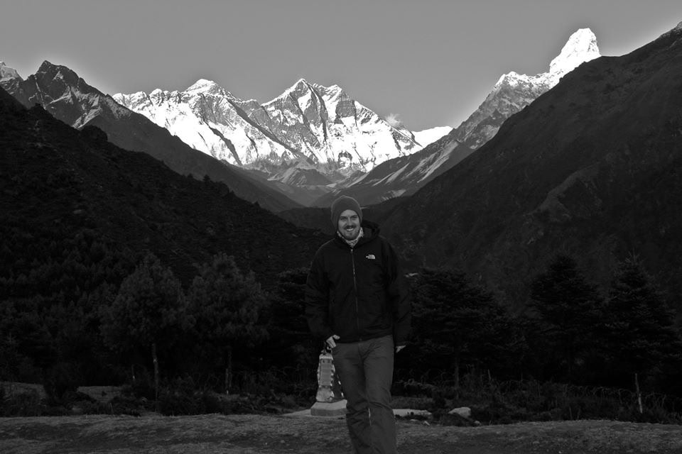 A person stands outdoors, smiling in front of a background of snow-capped mountains and trees in Nepal. The sky is clear, and the mountains are prominently visible. It's a moment perfect for any family photobook.