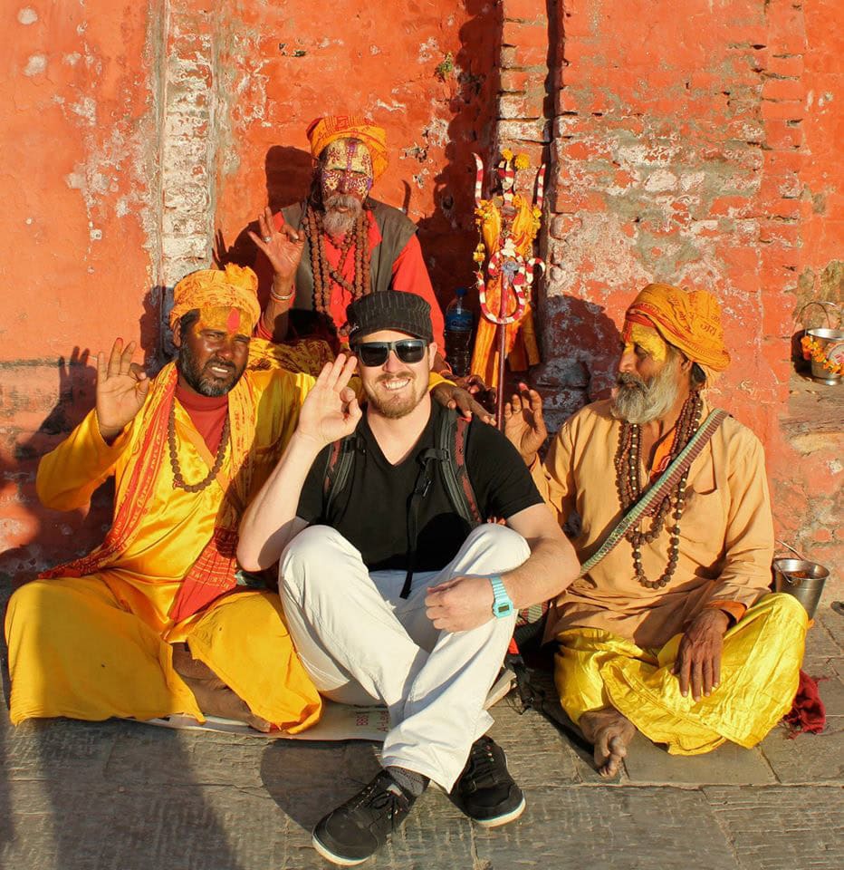 A person sits cross-legged and smiling with three men dressed in traditional attire near a weathered, red brick wall. All four individuals hold a hand up in a gesture, capturing a warm family moment that would belong perfectly in a photobook celebrating the rich culture of Nepal.