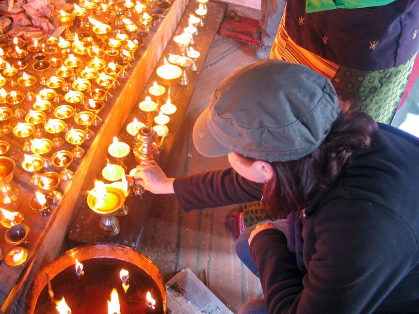 A person wearing a cap and jacket lights a butter lamp in front of a large display of lit lamps on a table, reminiscent of the vibrant cultural tapestry found in Nepal. Another individual in traditional clothing stands nearby, adding to the sense of exploration and discovery.