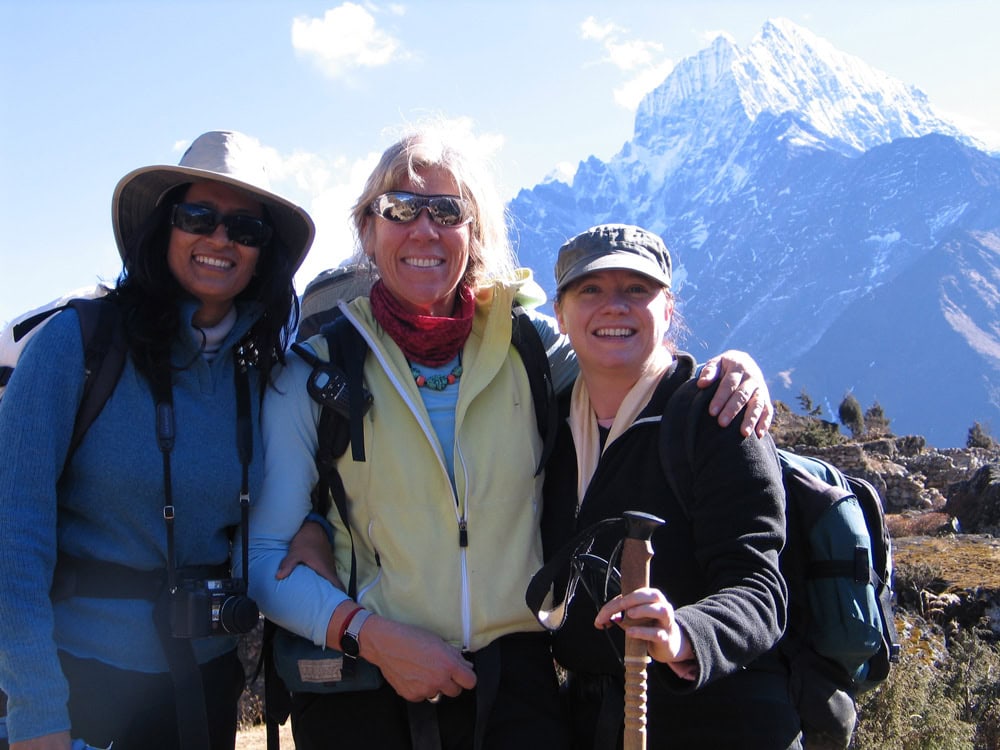 Three women in hiking gear, possibly on a family adventure, smile and pose for a photo with a snowy mountain peak in the background—a perfect moment for their travel photobook from Nepal.