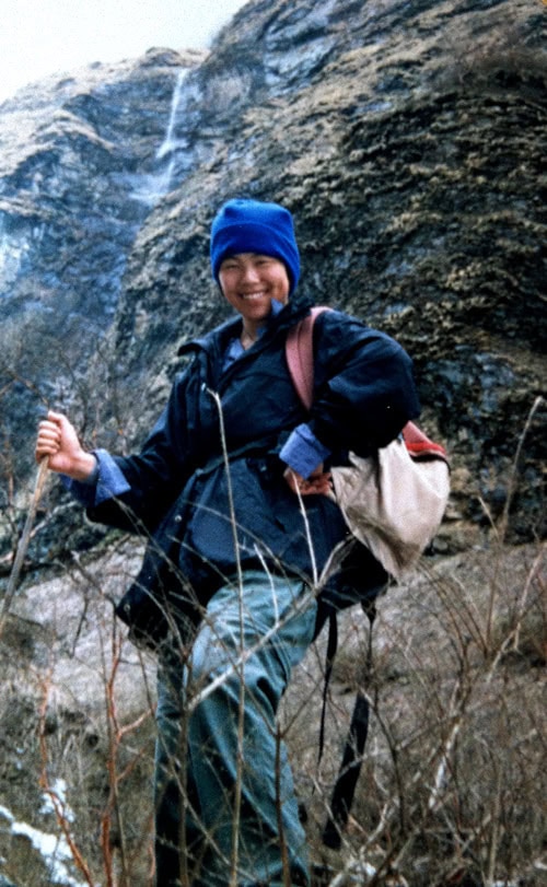 Person wearing outdoor gear and blue hat stands on a rocky, grassy hillside in Nepal with a trekking pole and a backpack, smiling at the camera, perfect for the family photobook.