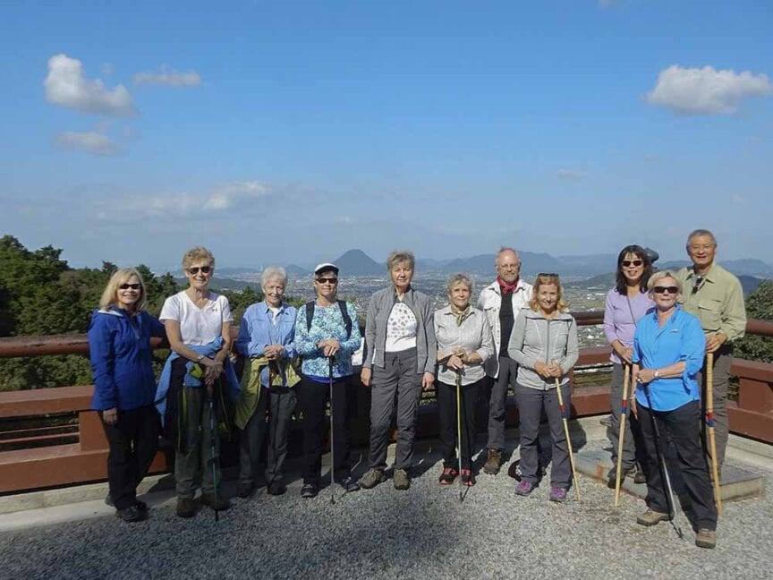 A group of ten people with hiking gear stands on a scenic lookout with mountains in the background on a clear day, reminiscent of the historic trails of Shikoku.