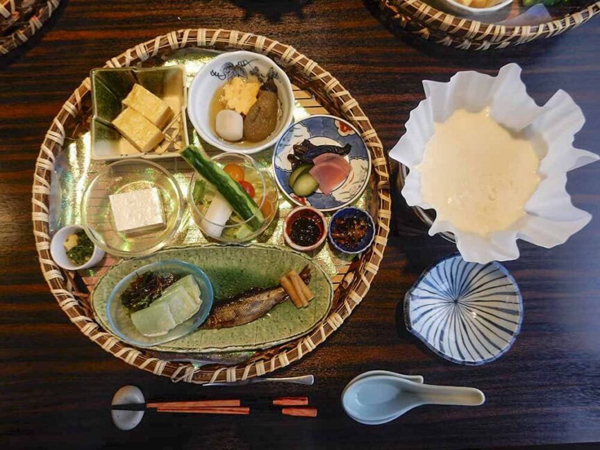 A traditional Japanese meal presentation with various small dishes including pickles, tofu, fish, vegetables, sauces, and a soup bowl on a dark wooden table—reminiscent of the culinary heritage found in Shikoku after a day of hiking its historic trails.