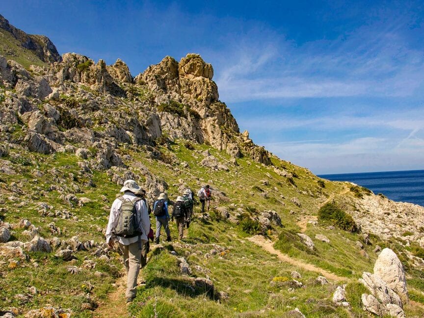 A group of hikers wearing backpacks walks along a rugged, grassy mountain trail with rocky formations under a clear blue sky, near a body of water—one of the many wondrous Europe travel experiences.