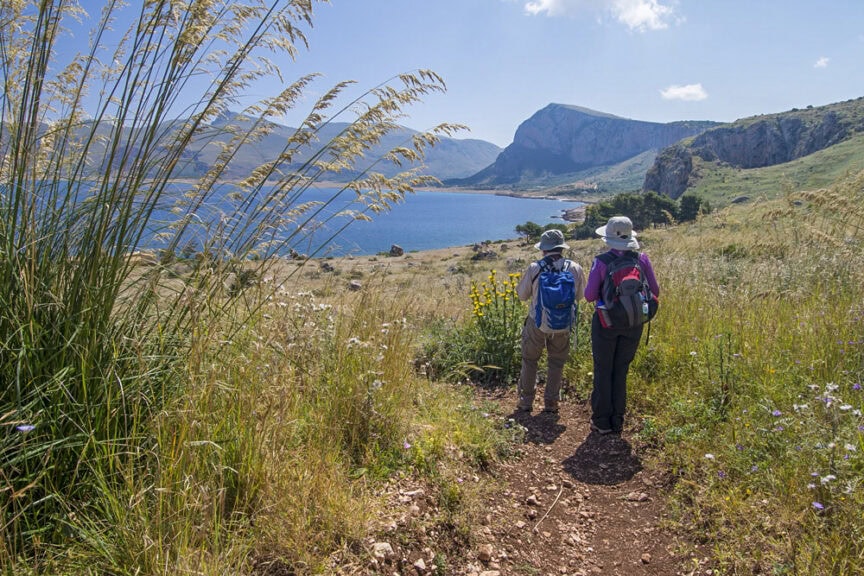 Two hikers with backpacks and hats walk along a scenic trail in Sicily, surrounded by grass, overlooking a shimmering body of water and distant mountains under the clear sky. The idyllic landscape seems to spotlight the raw beauty of nature.