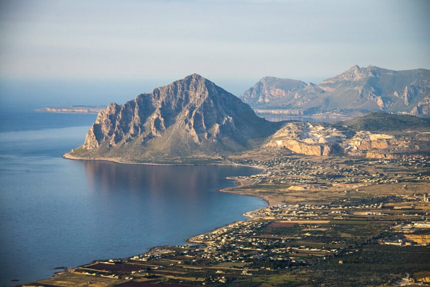 Aerial view of a coastal landscape in Sicily with a prominent mountain surrounded by farmland and buildings, meeting the calm sea under a clear sky—a perfect Spotlight for Sicily travel.