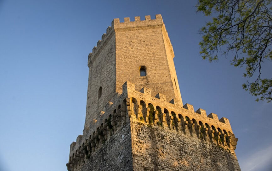A large stone tower with battlements rises against a clear blue sky, partially framed by a leafy tree branch on the right side, as if Sicily's historic architecture were under a natural spotlight.