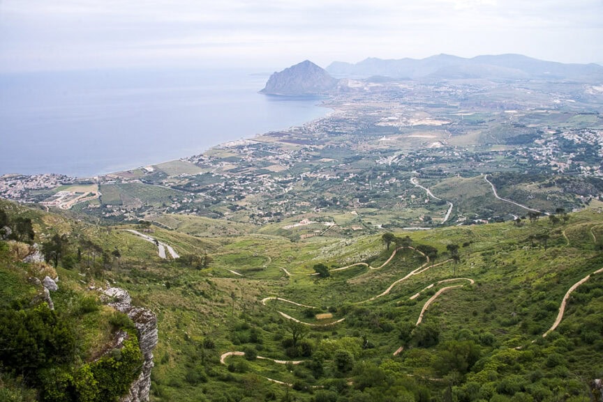 Aerial view of a coastal landscape in the spotlight: Sicily, featuring winding roads, lush green hills, a sprawling town, and a distant mountain near the sea.