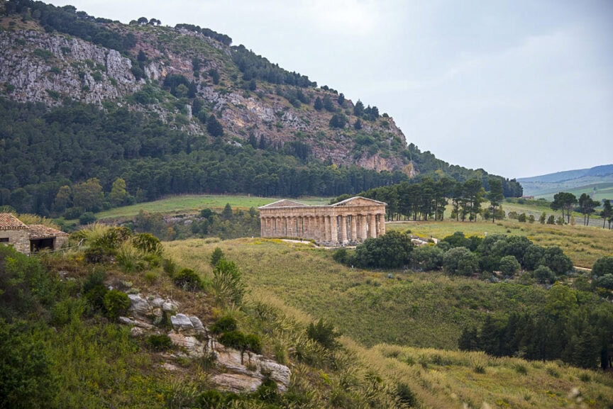 Set in the rolling hills of Sicily, this ancient Greek temple with its iconic columns stands majestically amid lush greenery and trees, all under a dramatic cloudy sky. The beautiful landscape puts the spotlight on this historic architectural gem.