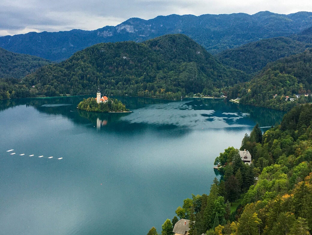 Aerial view of Lake Bled in Slovenia, featuring a small island with a church, surrounded by forested hills and mountains under a cloudy sky—an idyllic scene perfect for your next trip to Slovenia.
