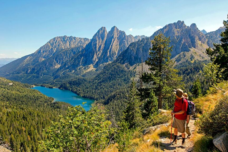 Two hikers stand on a trail overlooking a scenic view of a mountain range and a blue lake surrounded by dense forest, ready for their 9-day travel adventure.