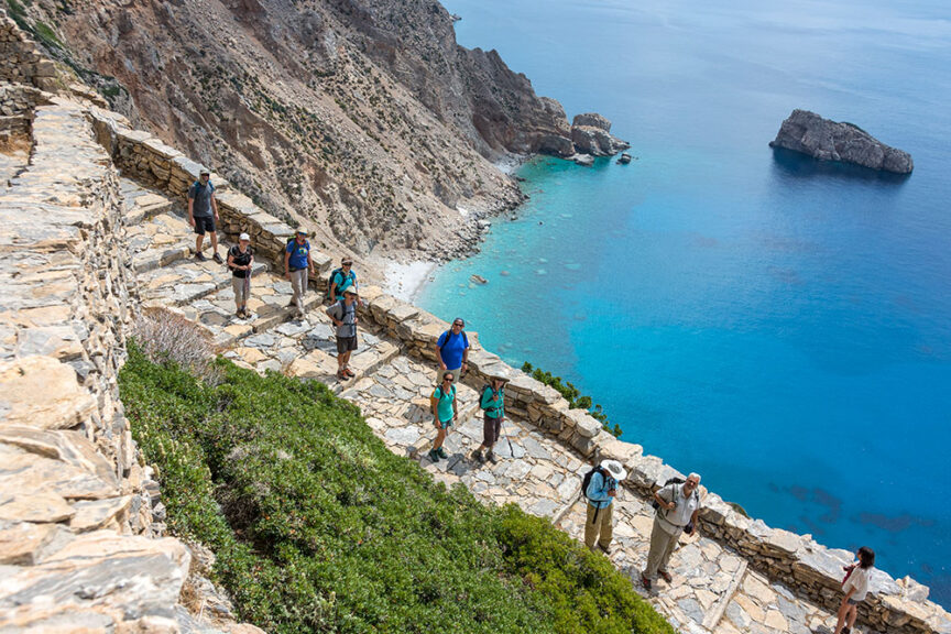 A group of hikers walks along a stone path on a steep cliffside in Greece, with a view of the blue ocean below and rocky outcrops in the distance.
