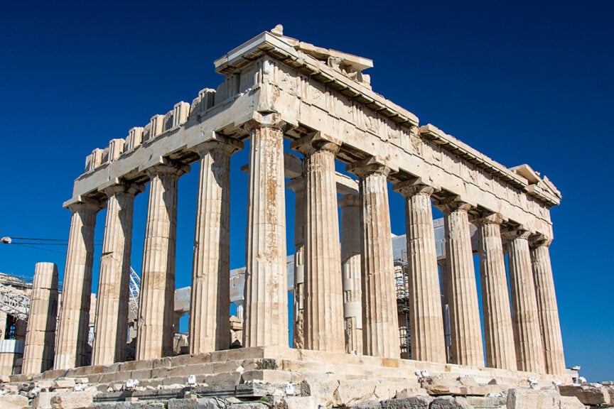 The image shows the Parthenon, an ancient Greek temple on the Acropolis in Athens, Greece, with its iconic columns and partially ruined structure against a clear blue sky. Surrounding trails add to the allure of this historic landmark.