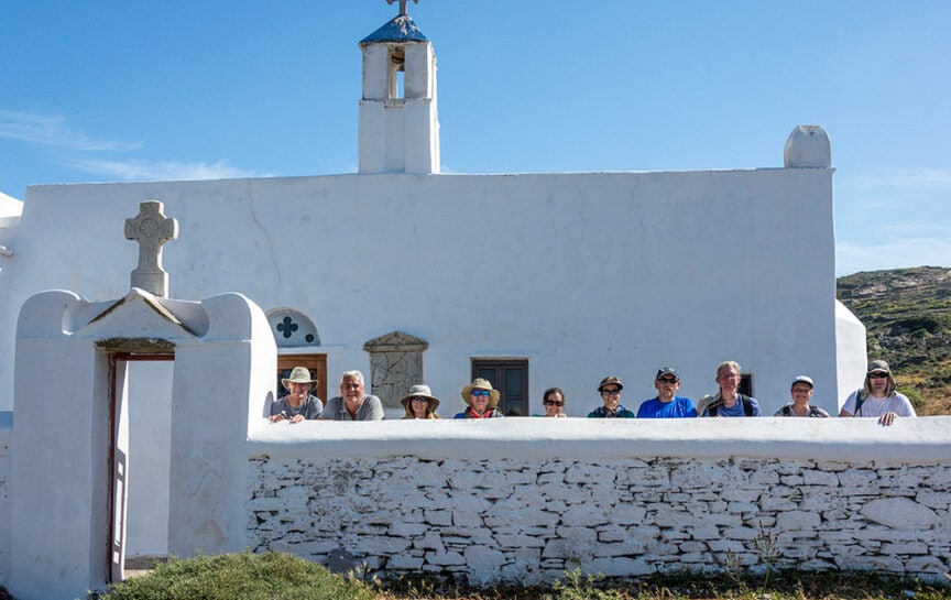 A group of people posed behind a white stone wall in front of a small white church with a cross on the gate, under a clear blue sky, capturing the serene beauty of Greece.