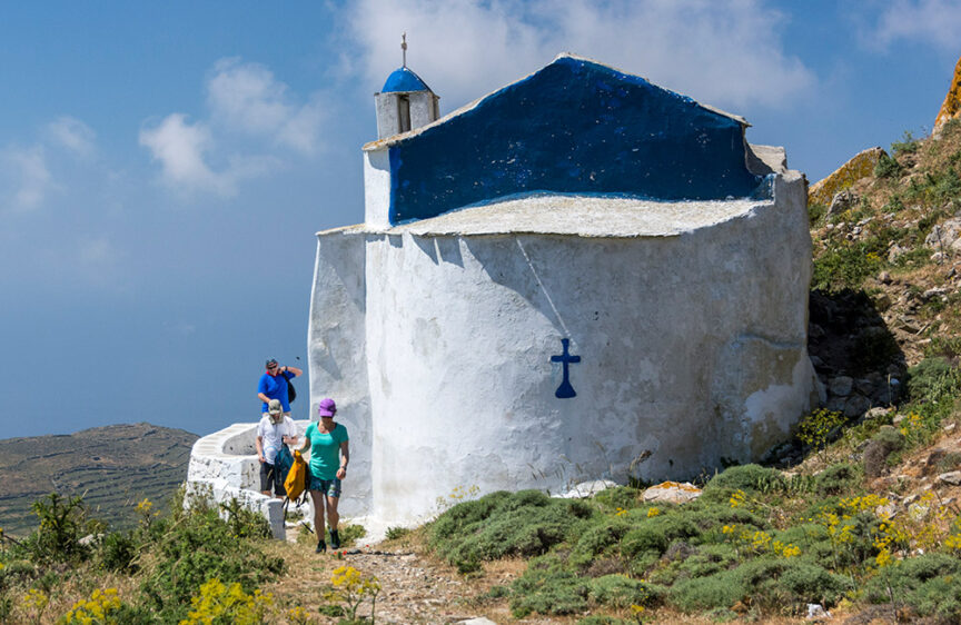 A group of people walking near a small white and blue chapel on a hillside in Greece, with greenery, flowers, and scenic trails around, all beneath a partly cloudy sky.