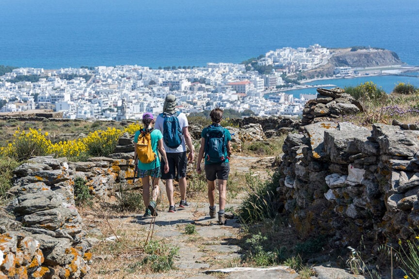 Three people with backpacks hike a stone path in Greece, with a scenic view of a coastal town and the sea in the distance, epitomizing the allure of travel along these historic trails.
