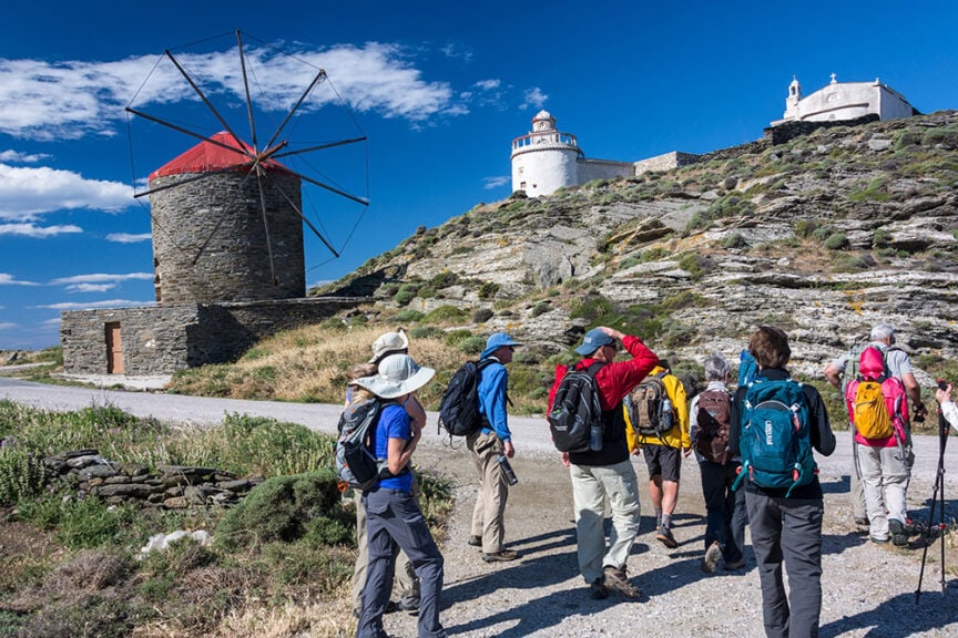 A group of people with backpacks and sun hats hike along scenic trails towards a windmill and a white building on a rocky hillside in Greece, all under a clear blue sky.