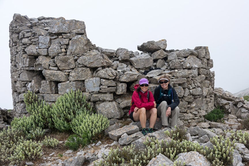 Two people wearing outdoor gear sit in front of a dilapidated stone structure, surrounded by small green shrubs and plants, under a foggy Greek sky, taking a break from their travel along the rugged trails.