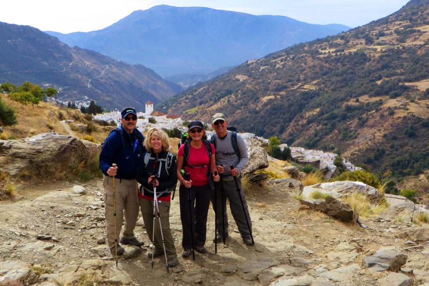 Four authentic hikers with trekking poles stand on a mountain trail in Spain, with a scenic valley and distant mountains in the background.