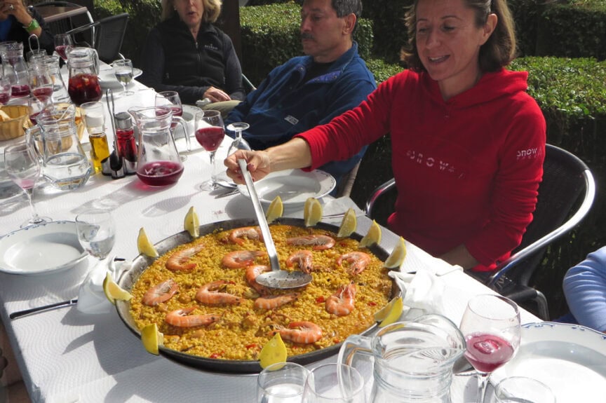 A woman authentically serves paella from a large pan at an outdoor table set with glasses, pitchers, and various dishes, surrounded by people, evoking the vibrant spirit of Spain.