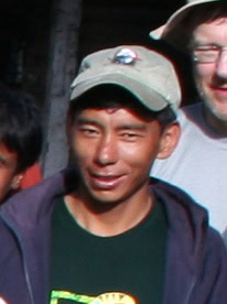 A person wearing a baseball cap and dark clothing stands outdoors in Nepal, smiling, as if posing for a photobook.