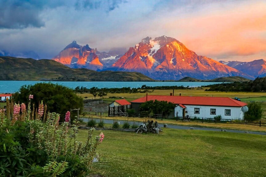 A rural landscape features a house with a red roof and green fields in the foreground. In the background, a mountain range, reminiscent of Patagonia, is bathed in sunset light, complemented by a serene lake—ideal for epic hikes.