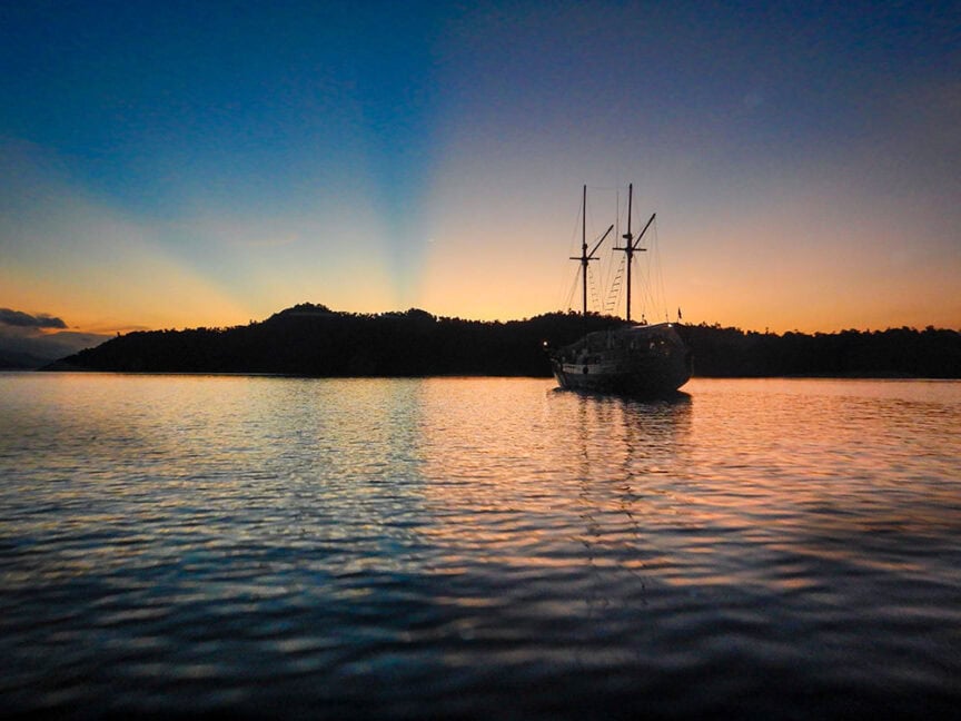 A sailboat is anchored on the water's surface with the silhouette of an island in the background during sunset, casting a gradient of orange to blue in the sky, evoking dreams of exploring places like Raja Ampat.