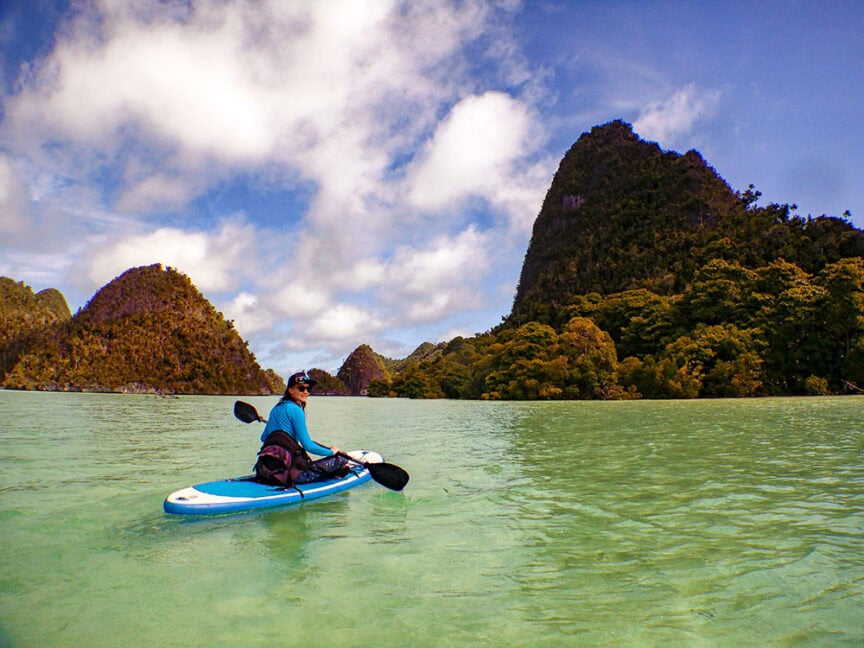 A person kayaks on a clear, turquoise body of water's surface surrounded by lush, green hills under a partly cloudy sky, exploring the natural beauty of Raja Ampat.