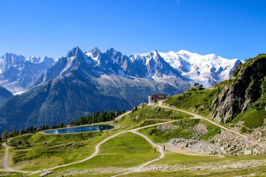 A scenic mountain landscape with rugged peaks, a grassy hillside, small buildings, and a pond under a clear blue sky. Winding paths are visible throughout the area, reminiscent of Trail Photos from the Tour du Mont Blanc.