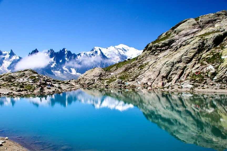 A clear turquoise lake reflects snow-capped mountains and a blue sky, surrounded by rocky shorelines and jagged peaks, offering perfect moments for Trail Photos along the Tour du Mont Blanc.