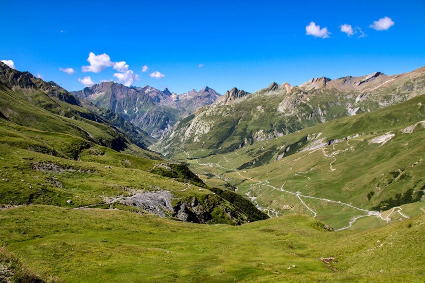 A panoramic view of the Tour du Mont Blanc reveals a mountainous landscape with green slopes, winding trails, and a clear blue sky adorned with scattered clouds. Trail photos simply can't capture its full majesty.