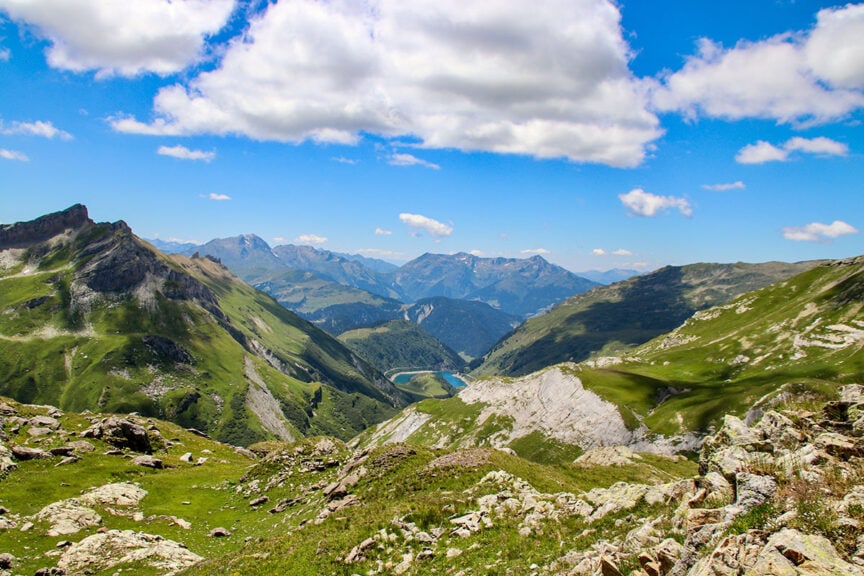 A panoramic view of the Tour du Mont Blanc with rocky peaks, lush green valleys, and a blue lake under a sky with scattered clouds—perfect for capturing stunning trail photos.