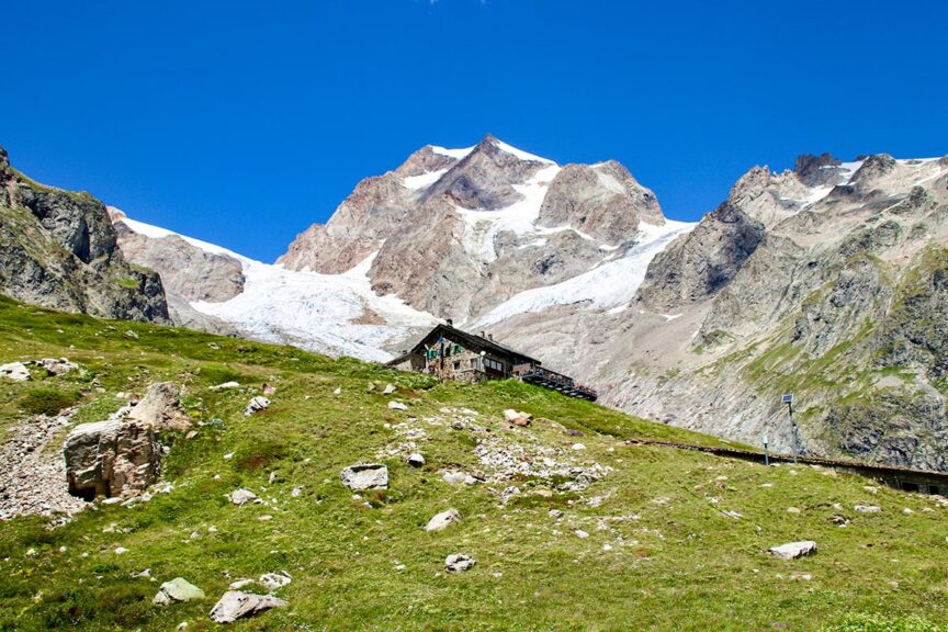 A small, rustic house sits on a grassy hill beneath towering, snow-capped mountains on a clear day, perfect for capturing memorable trail photos during your Tour du Mont Blanc adventure.