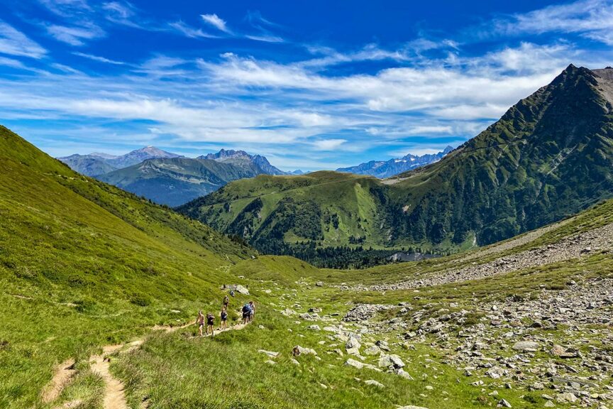 A group of people hike along a trail in a lush green valley surrounded by mountains under a partly cloudy blue sky, capturing perfect Trail Photos reminiscent of the stunning Tour du Mont Blanc.