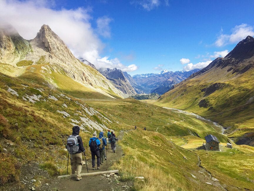 Hikers with backpacks trek along a mountain trail under a blue sky with scattered clouds, surrounded by green and rocky terrain. A small building is visible in the distance near a winding river, perfect for those considering a 9-day adventure.