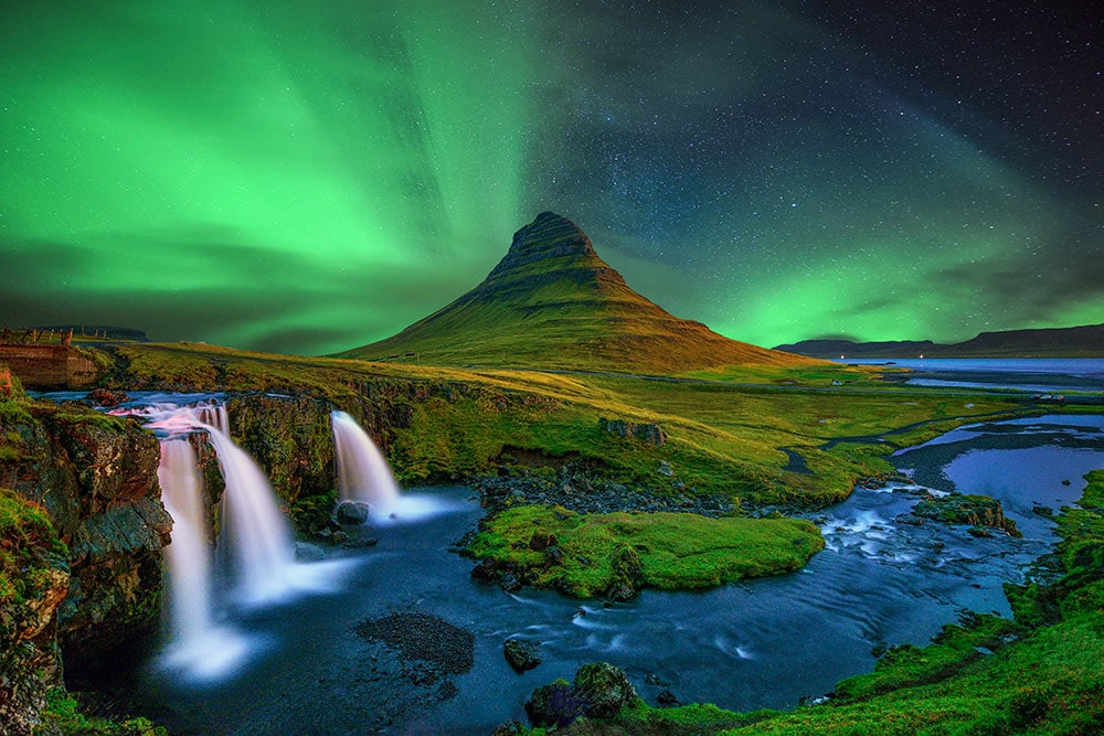 Northern lights illuminate the sky above Kirkjufell mountain and waterfall in Iceland, one of the best places for aurora adventures, against a backdrop of grassy landscapes and flowing water.