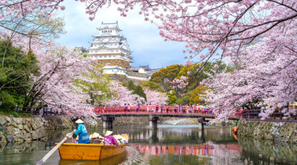 A wooden boat with people in traditional hats on a river surrounded by cherry blossoms, with a white castle and a red bridge in the background captures the essence of festivals celebrated around the world.