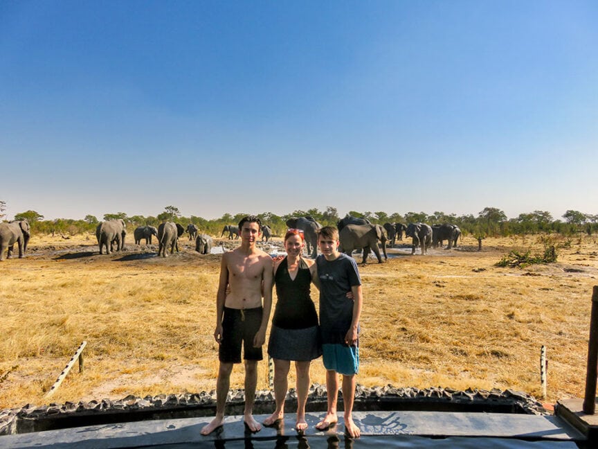 Three people standing on a platform in a dry, grassy area with a herd of elephants in the background under a clear blue sky, capturing the essence of the Great Elephant Migration in Botswana.